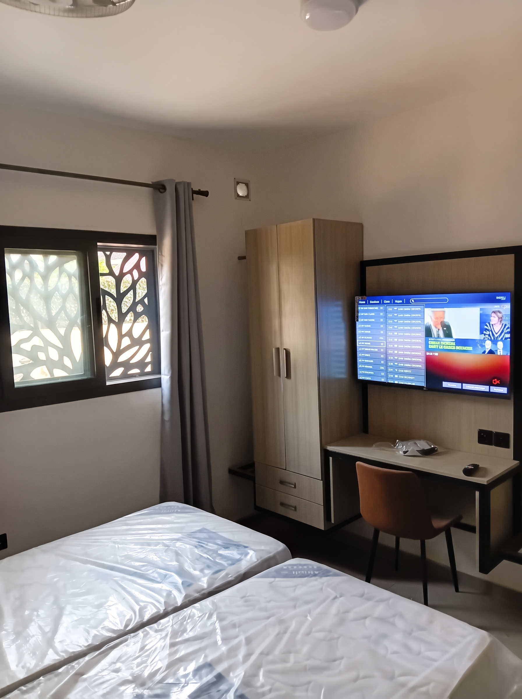 Chambre de la résidence avec literie, armoire en bois clair, bureau et téléviseur mural