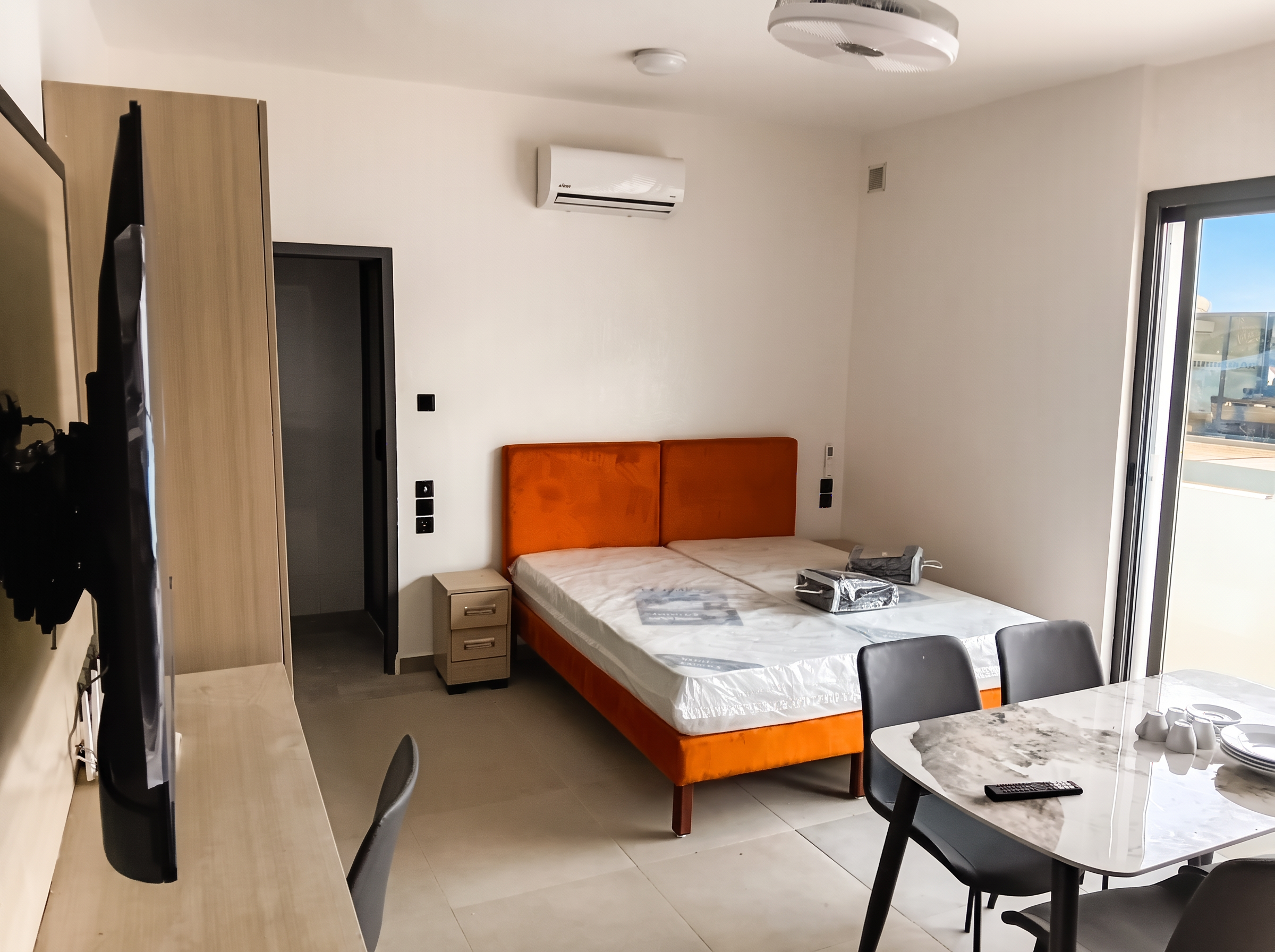 Chambre lumineuse avec lit, climatiseur mural, ventilateur de plafond, table et baie vitrée